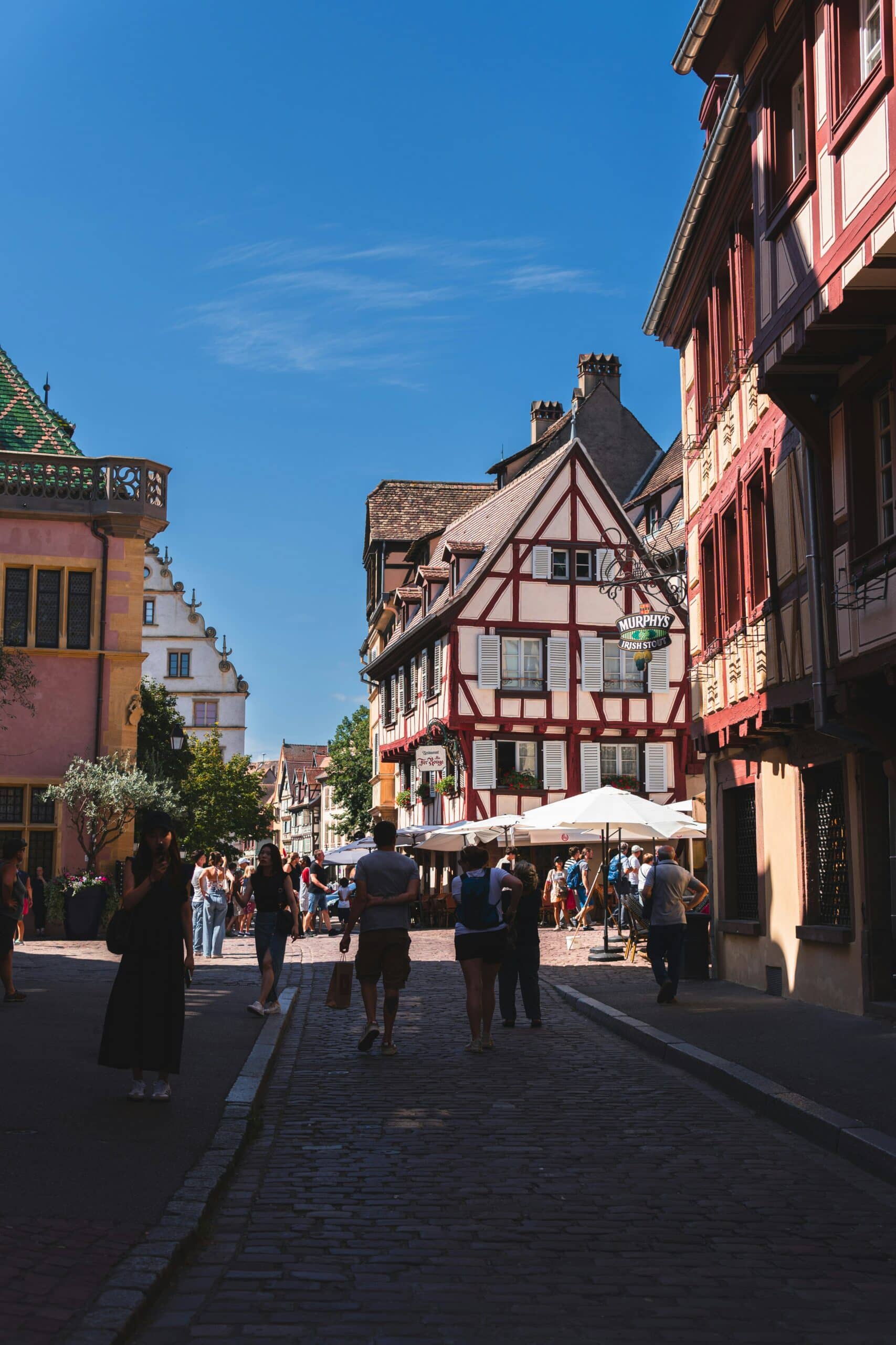 Rue animée d’un village alsacien avec maisons à colombages, terrasses et passants