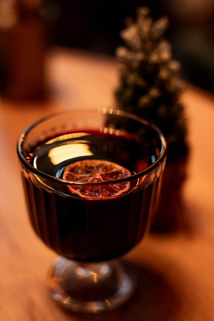 Verre de vin chaud aux épices avec une tranche d’orange séchée, servi sur une table en bois dans une ambiance hivernale chaleureuse.